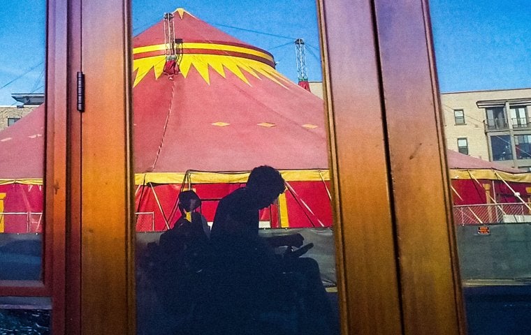 Image of a reflection in a window. A man in a wheelchair is looking downwards and holding a camera. In the background is a giant red circus tent.
