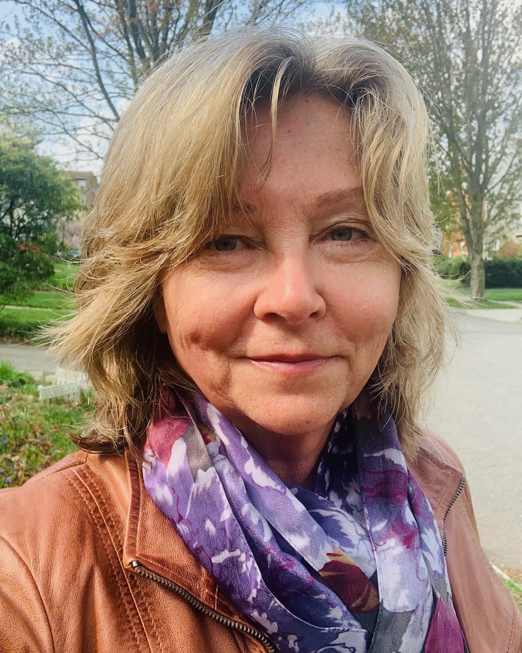 Image of a White woman with blonde hair wearing a purple scarf and rust colored jacket. She looks contentedly towards the camera with a field of green grass behind her.