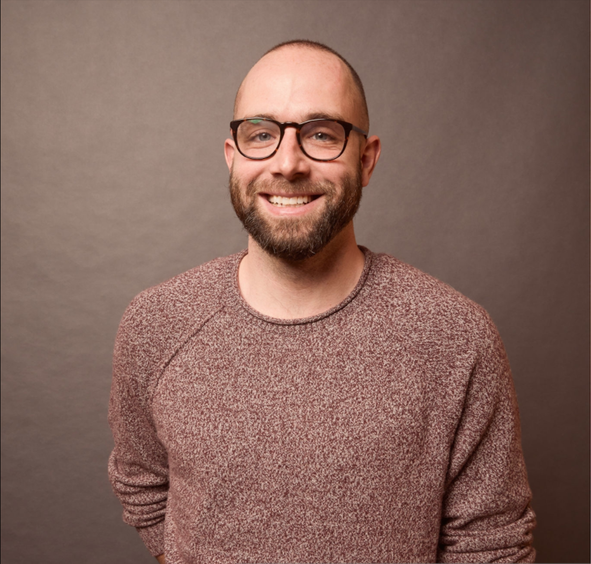 A man with a shaved head and beard, wearing glasses, smiles at the camera