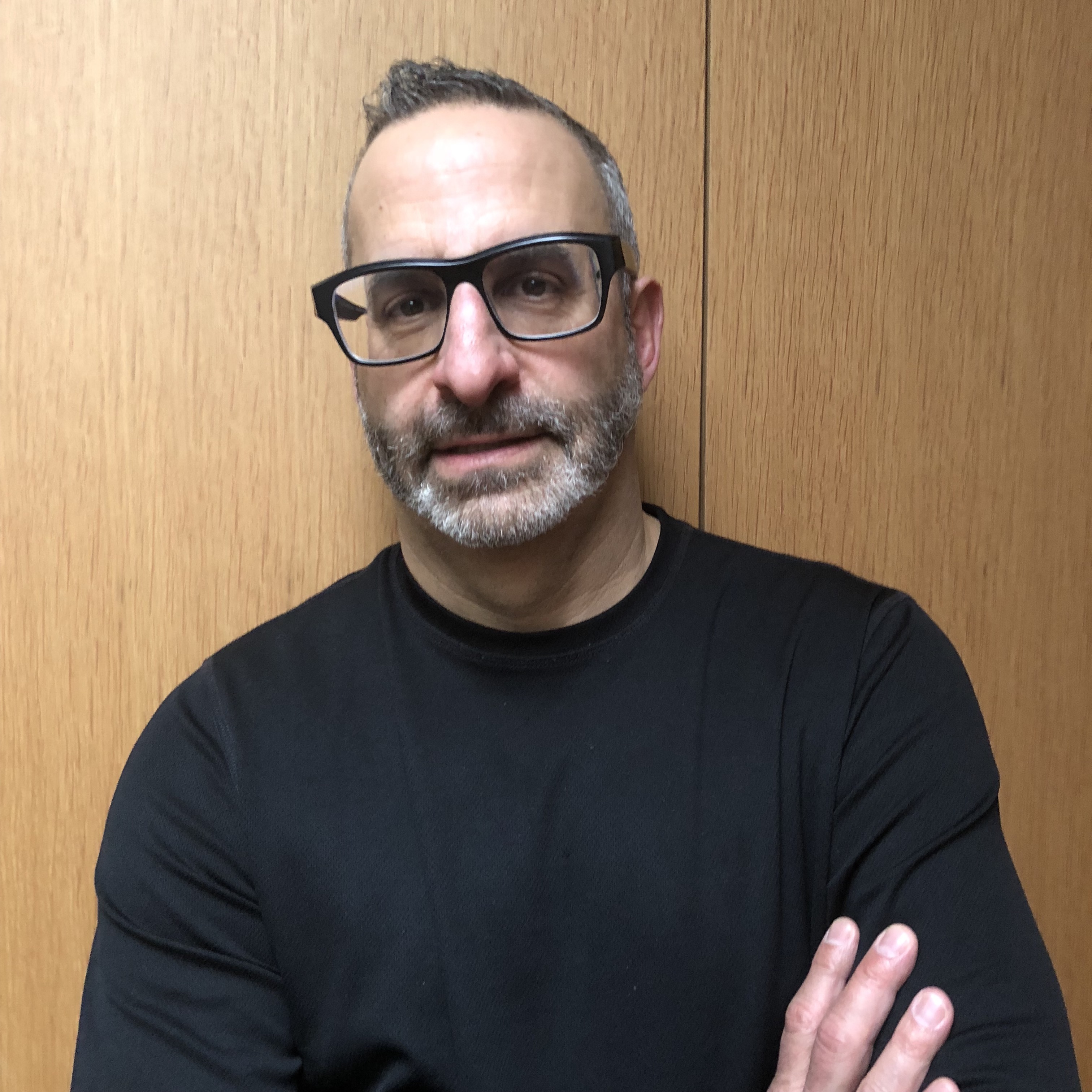 Image of white man with gray beard and black glasses with folded arms.