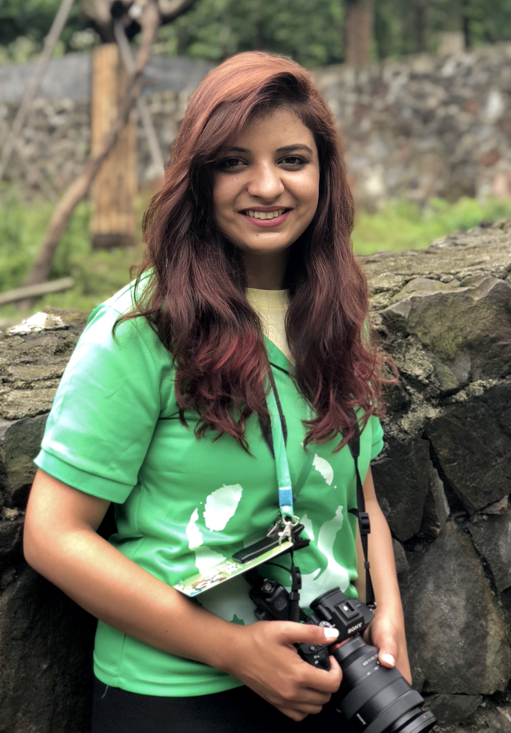 Women with long brown hair and a green t-shirt looks at the camera, smiling. She is holding a digital camera.