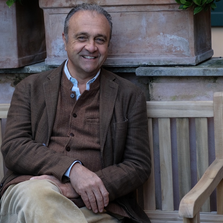 Image of a smiling man in a brown jacket with his arms folded on his lap and white collared shirt. Italian man with graying hair. 