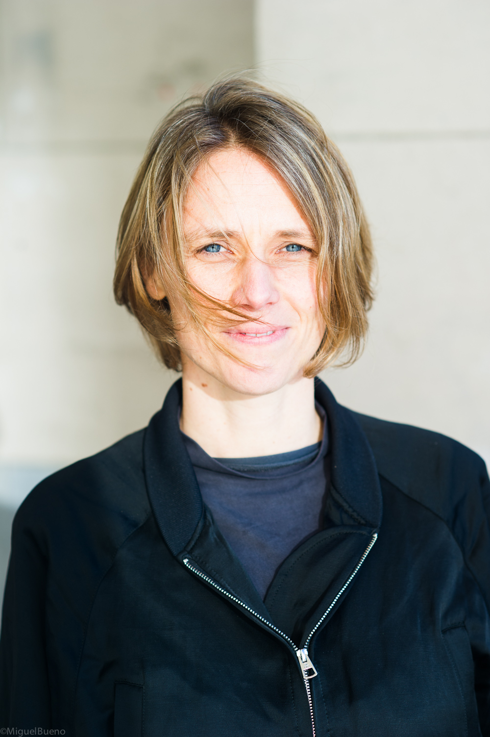 Image of White woman with short blonde hair slightly blowing in wind in front f her face. Wearing a black blazer and blue shirt in front of a gray wall. 