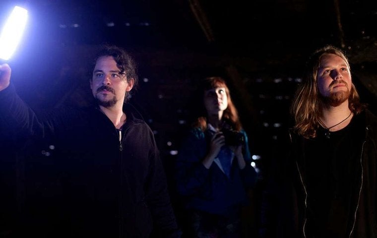 Three people stand in a dark room, one person on left holds up a light.