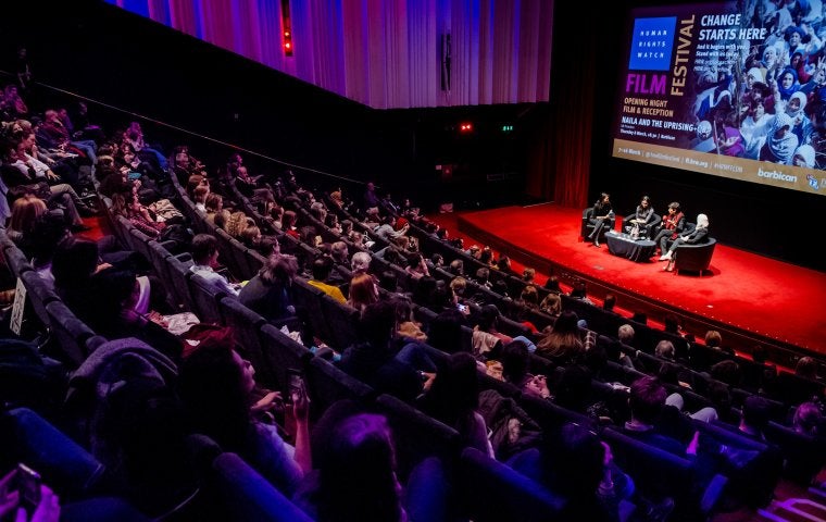 Image of opening night at the Film Festival at the Barbican in London. Audience clapping while speakers sit on red stage.