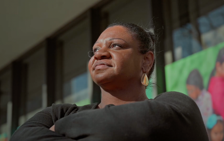 Photo of Black woman with hair pulled back in a bun, looking confidently up towards the light in left of image, with arms folded. 