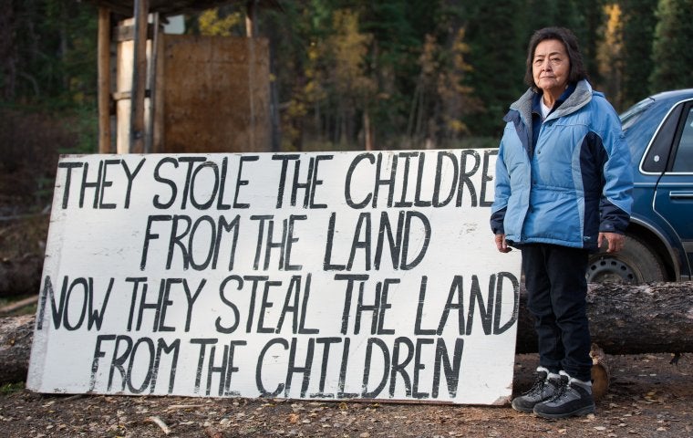 Photo of Elder by sign saying "They stole the children from the land, they steal the land from the children"