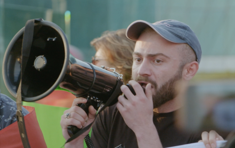 Image of white person with a megaphone in front of a crowd. 