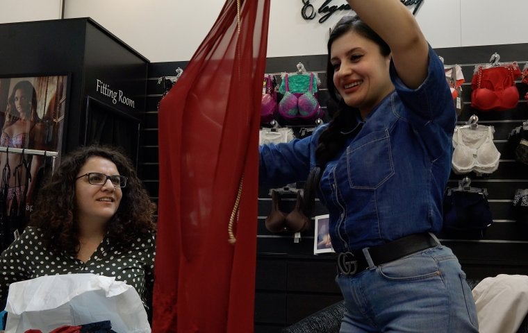 Photo of women in a lingerie shop. One is holding up a red piece of sheer fabric, and both women are smiling. 