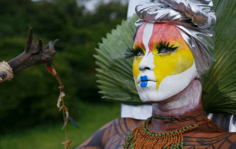 Image of non-binary person with white pink and yellow face makeup, blue lips and long eyelashes. Wearing a head wrap and holding a staff made of wood.