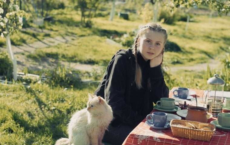Image of white girl with blonde hair pulled back into braids. She sits at a table outdoors with her cat. Red tablecloth on table and teacups. 