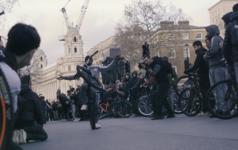Photo of a crowd of people in a circle on a London street, watching a young Black man doing a trick with his bike.