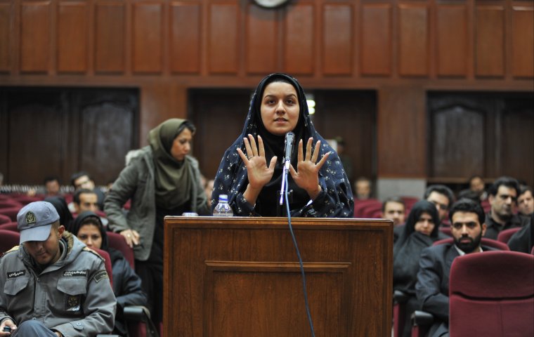 Photo of woman in court wearing hijab. She holds her hands up as she is speaking at a podium