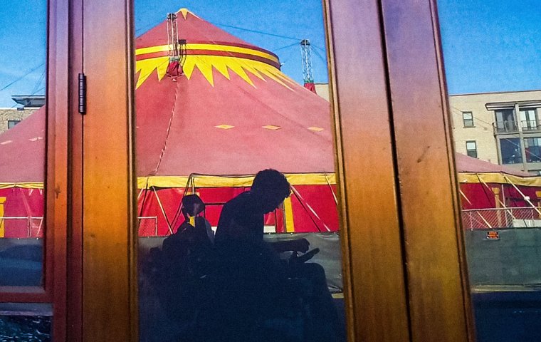 Image of a reflection in a window. A man in a wheelchair is looking downwards and holding a camera. In the background is a giant red circus tent.