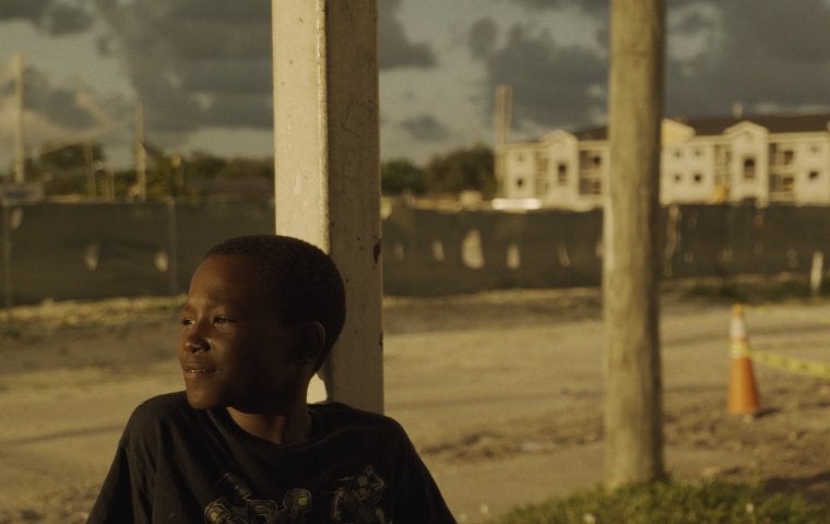 Photo of a young Black boy with short hair smiling and looking to the right. Behind him a housing structure sits, at the hour just before sunset.