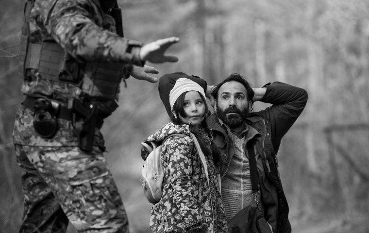 Black & white photo of barbed wire and a man on his knees with hands behind his head besides a young girl as someone in army fatigues holds out their hands