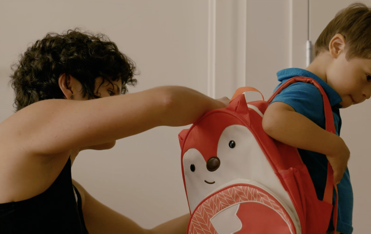 A young boy is facing sideways to the camera, an adult behind him is helping him pack his backpack which has a cartoon animal on it.