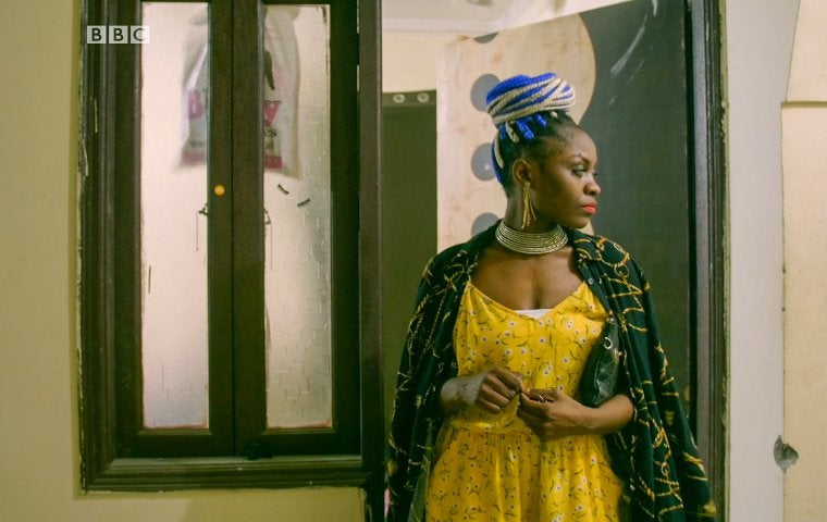 A young looking Black woman in a yellow dress and purple & white braids wrapped on top of her head stands in front of doors with glass panes, looking to her side. She is wearing a large gold necklace and long gold earrings.