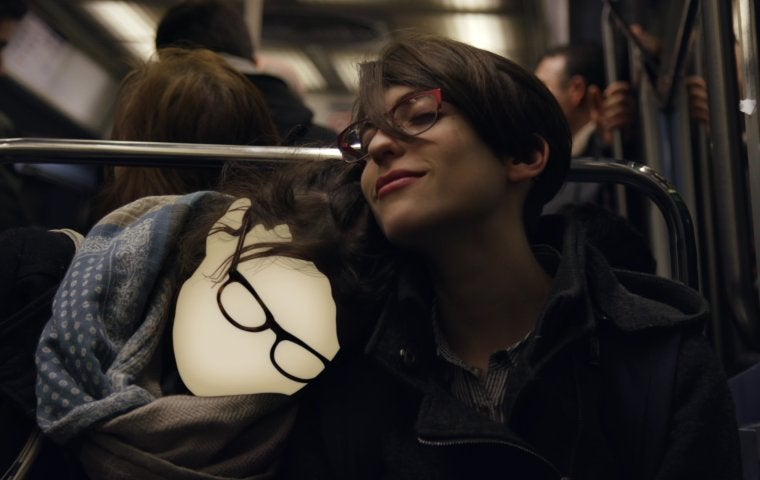 A young, white person with glasses and short dark hair sits on a train/bus looking up to their right. A computer-generated mask is on a 2nd person with glasses, who is leaning their head on the 1st person's right-hand side.