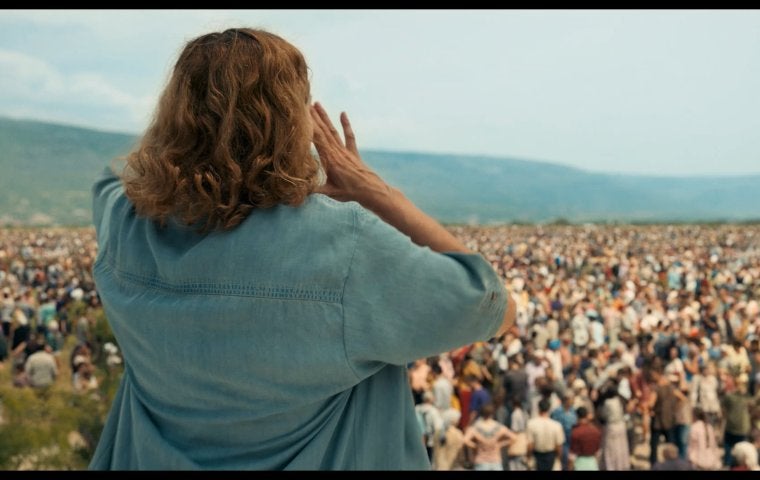 A crowd of people face a woman, we see the back of her head as she appears to be talking to them, hands cupped around her mouth to amplify her voice