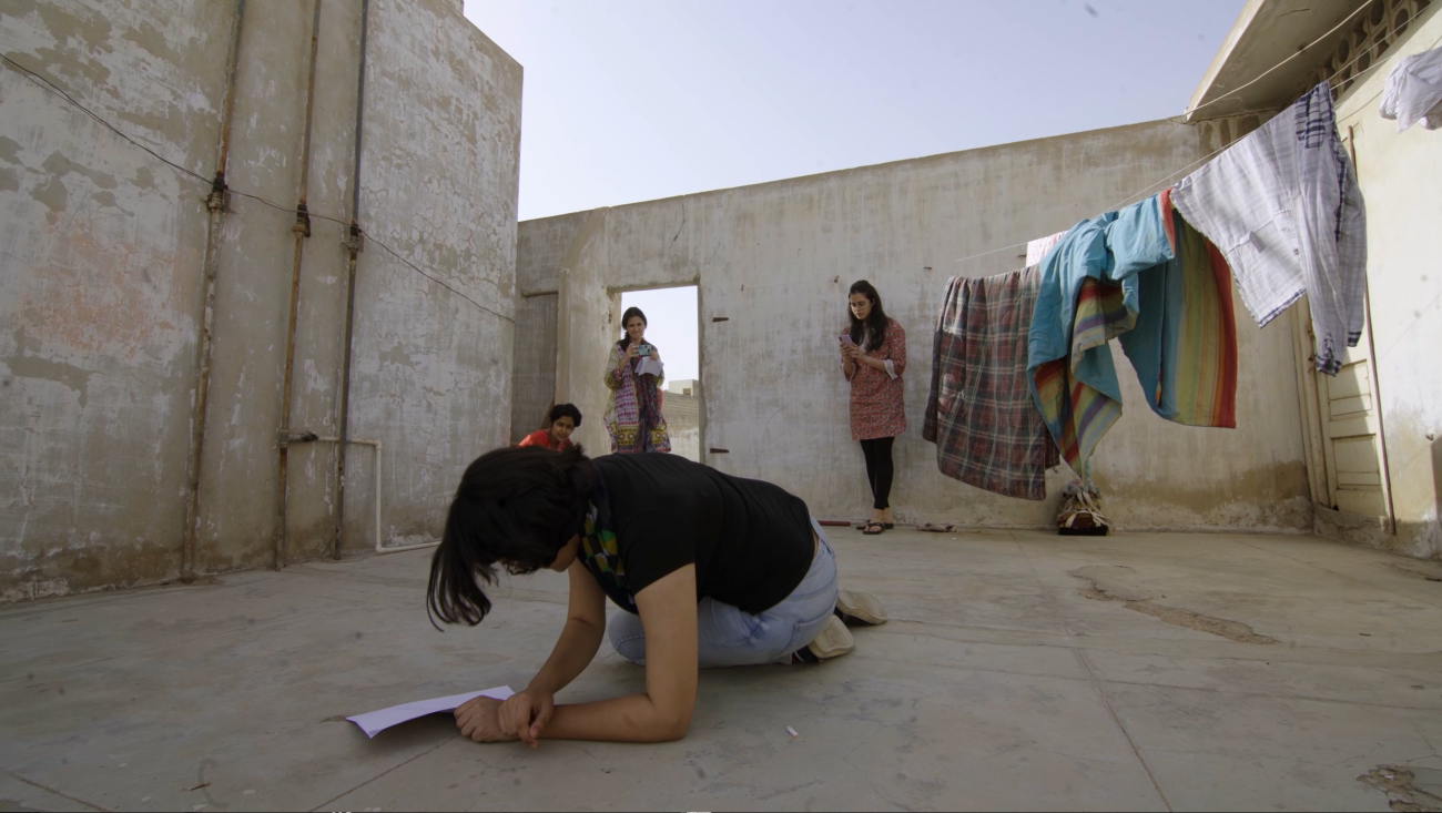 Woman lays on floor writing down notes as women stand behind her preparing for a performance.