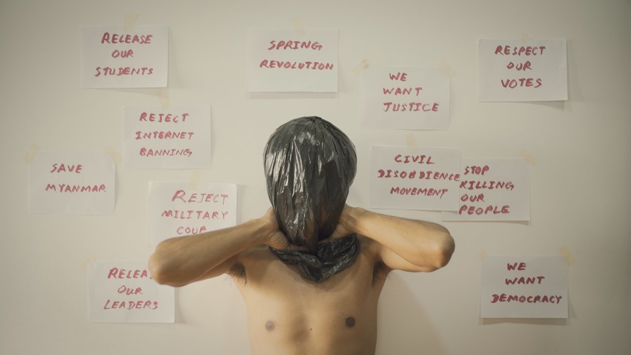 Man with a plastic bag over his head, signs behind him pinned to a wall read "We want justice" "Free the students" "Reject Military Rule" 