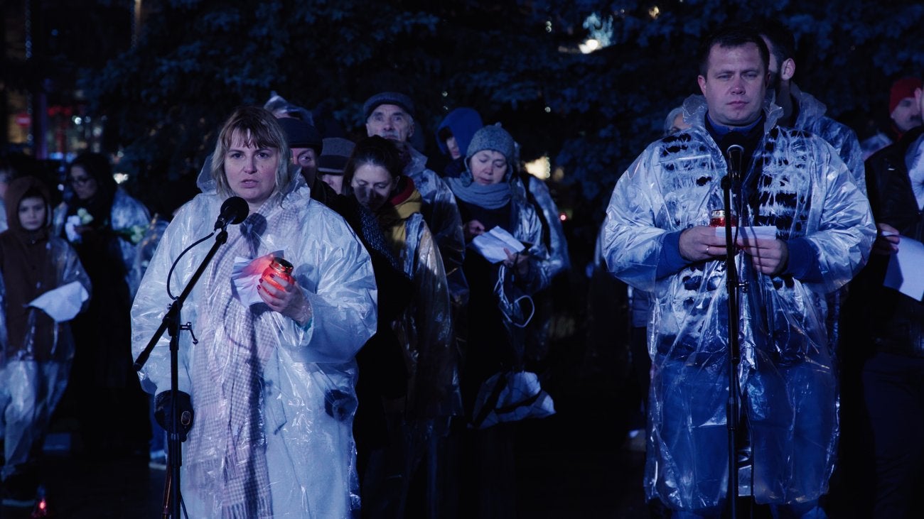 Photo of two Russian parents hosting a vigil on behalf of their imprisoned teenage daughter. Wearing rain ponchos and holding candles. 