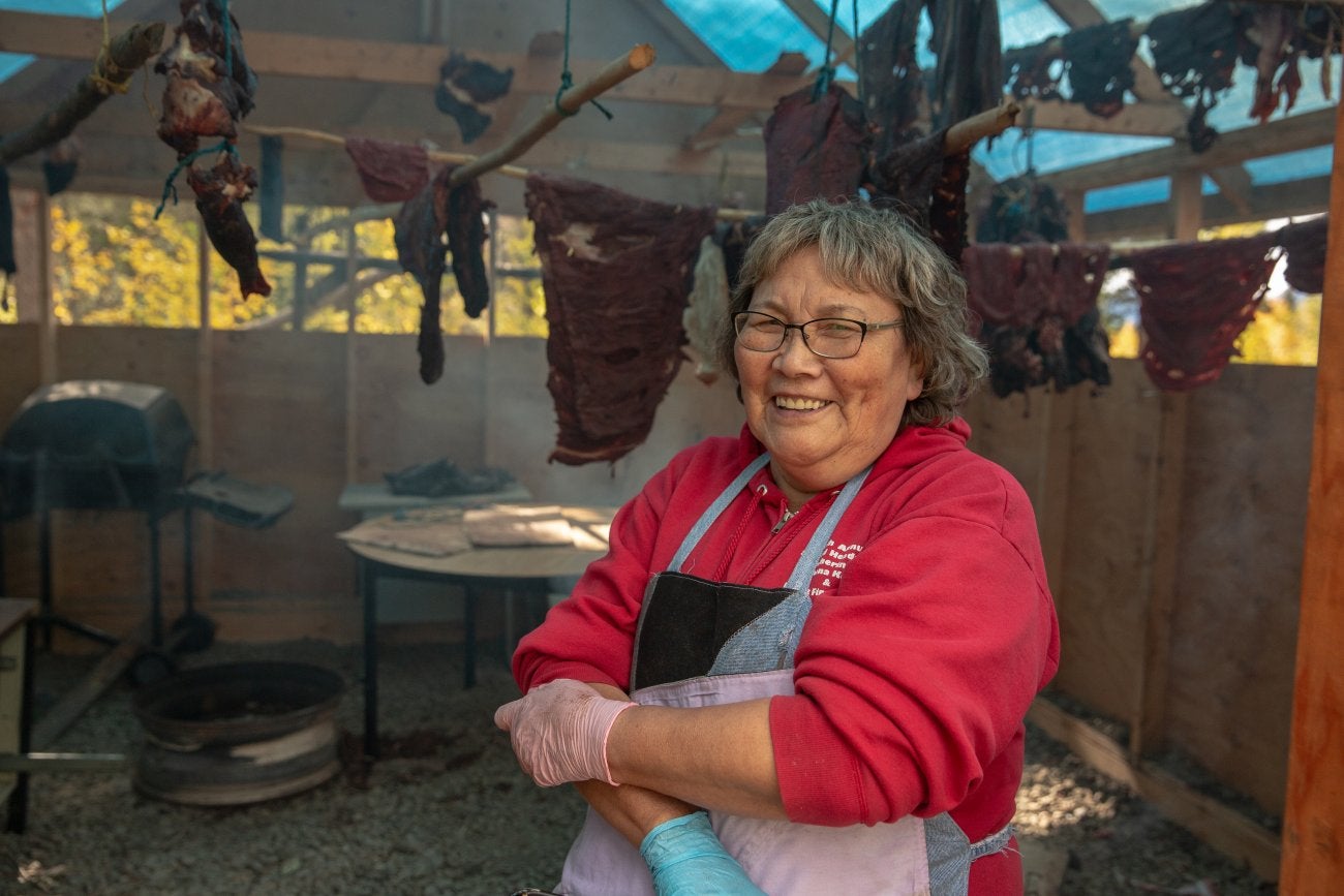 Image of Indigenous woman smiling with short gray hair, red shirt and apron