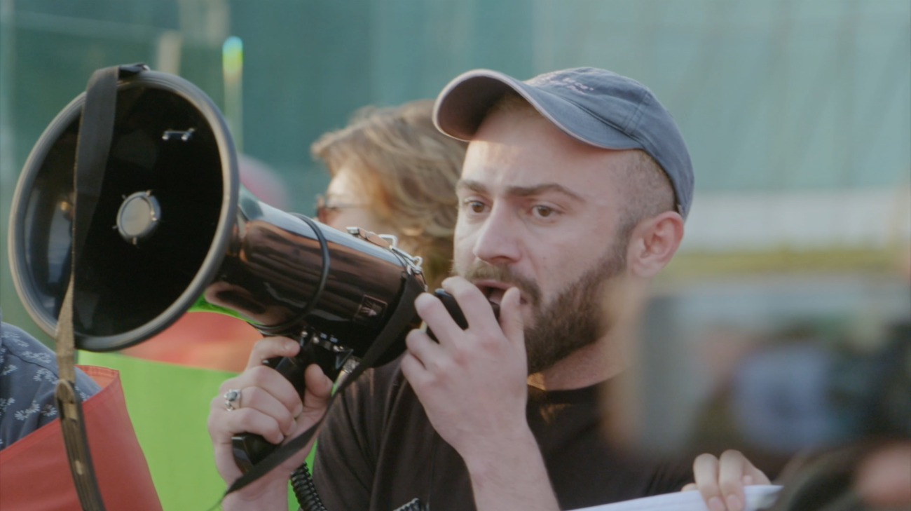 Image of white person with a megaphone in front of a crowd. 