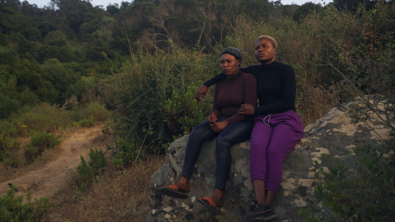 Image of two young African women sitting on a rock, one with an arm around the other. 