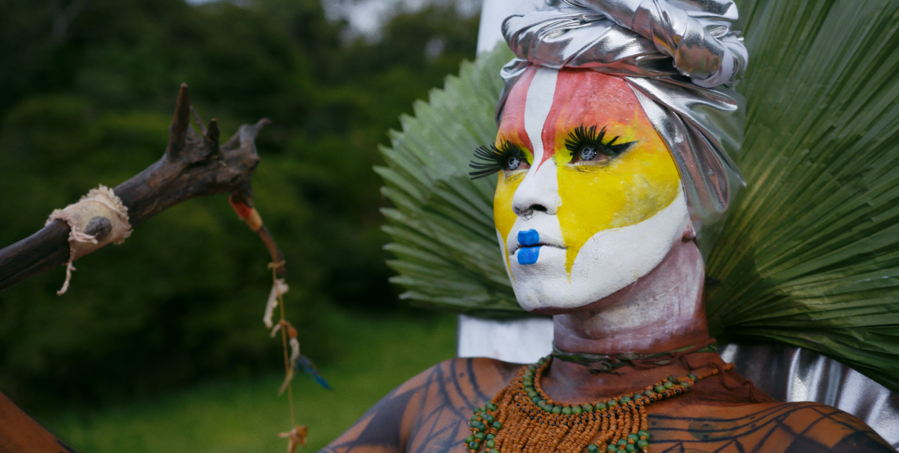 Image of non-binary person with white pink and yellow face makeup, blue lips and long eyelashes. Wearing a head wrap and holding a staff made of wood.
