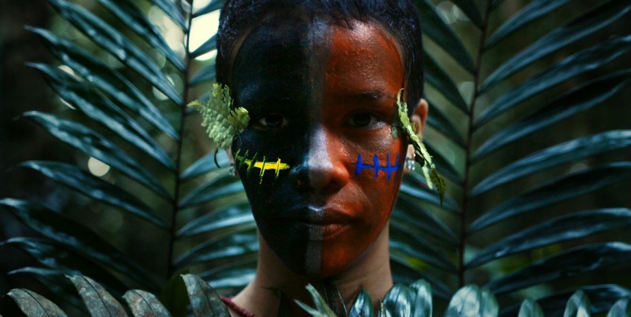 Image of a person looking at camera. Half of their face painted red with blue lines, half painted black with yellow lines. Background has palm tree leaves.
