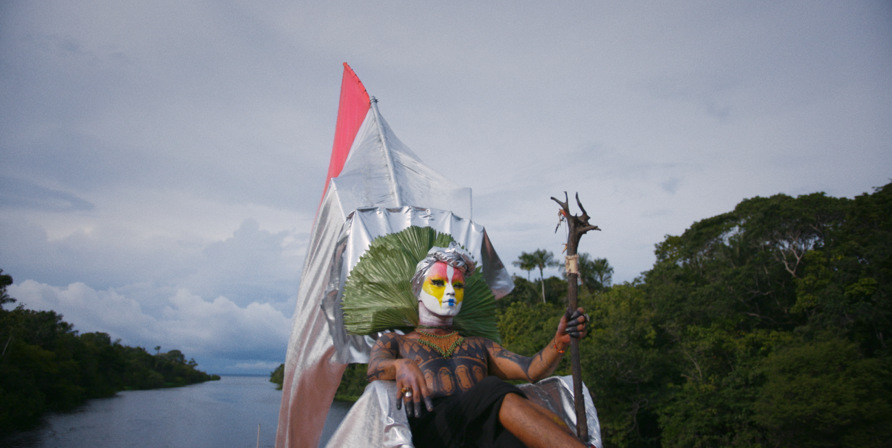 Photo of non-binary person sitting on throne made of leaves with flags. White makeup on their face with pink and green accents. Person is holding a wooden staff.