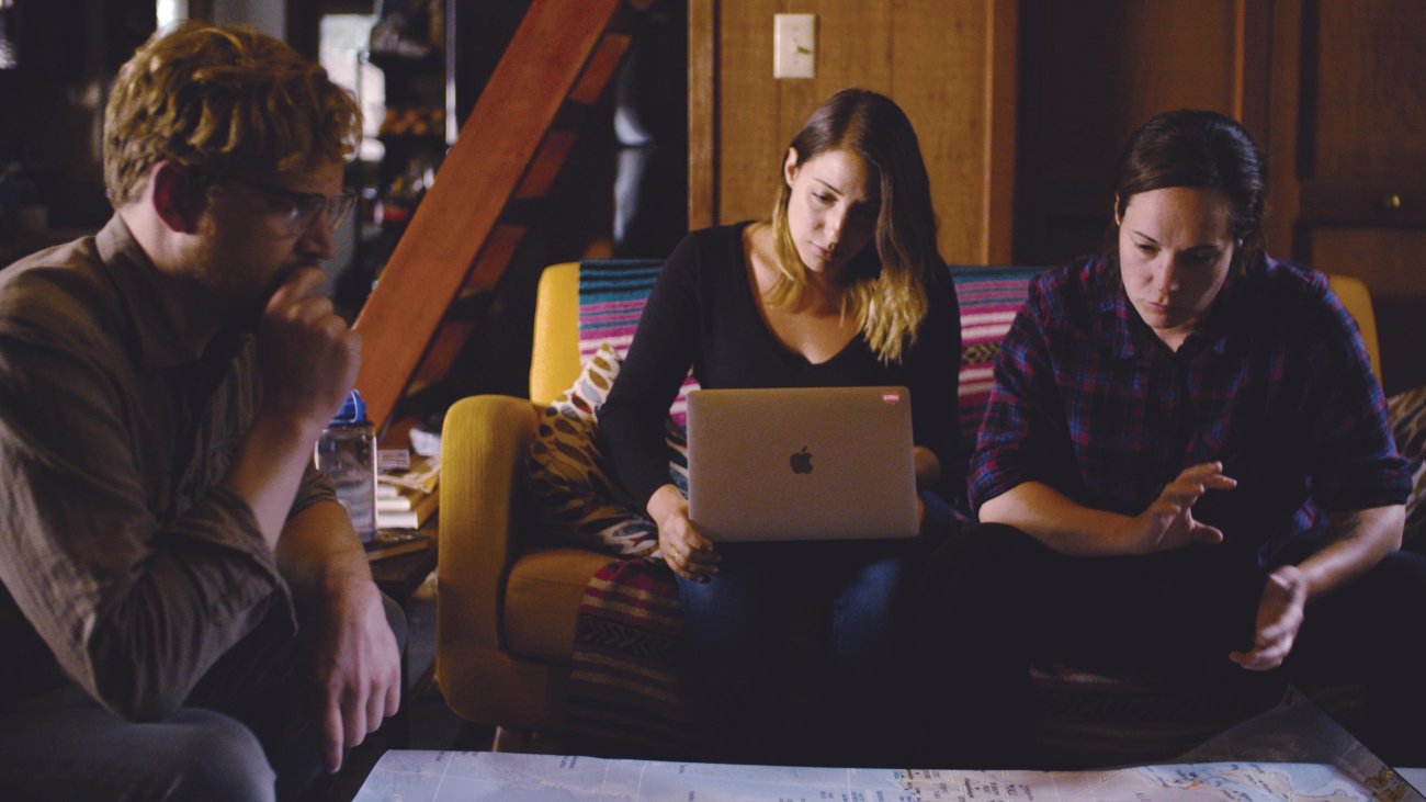 Three people sit around a low table with a large sheet of paper looking intently, while the central figure, a woman, looks down at her Mac Laptop 