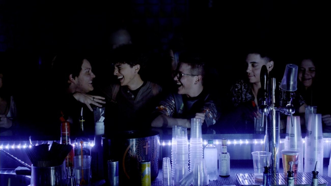 Image of four young Italian men at a bar smiling and laughing with each other. The bar has blueish lighting and background is dark.