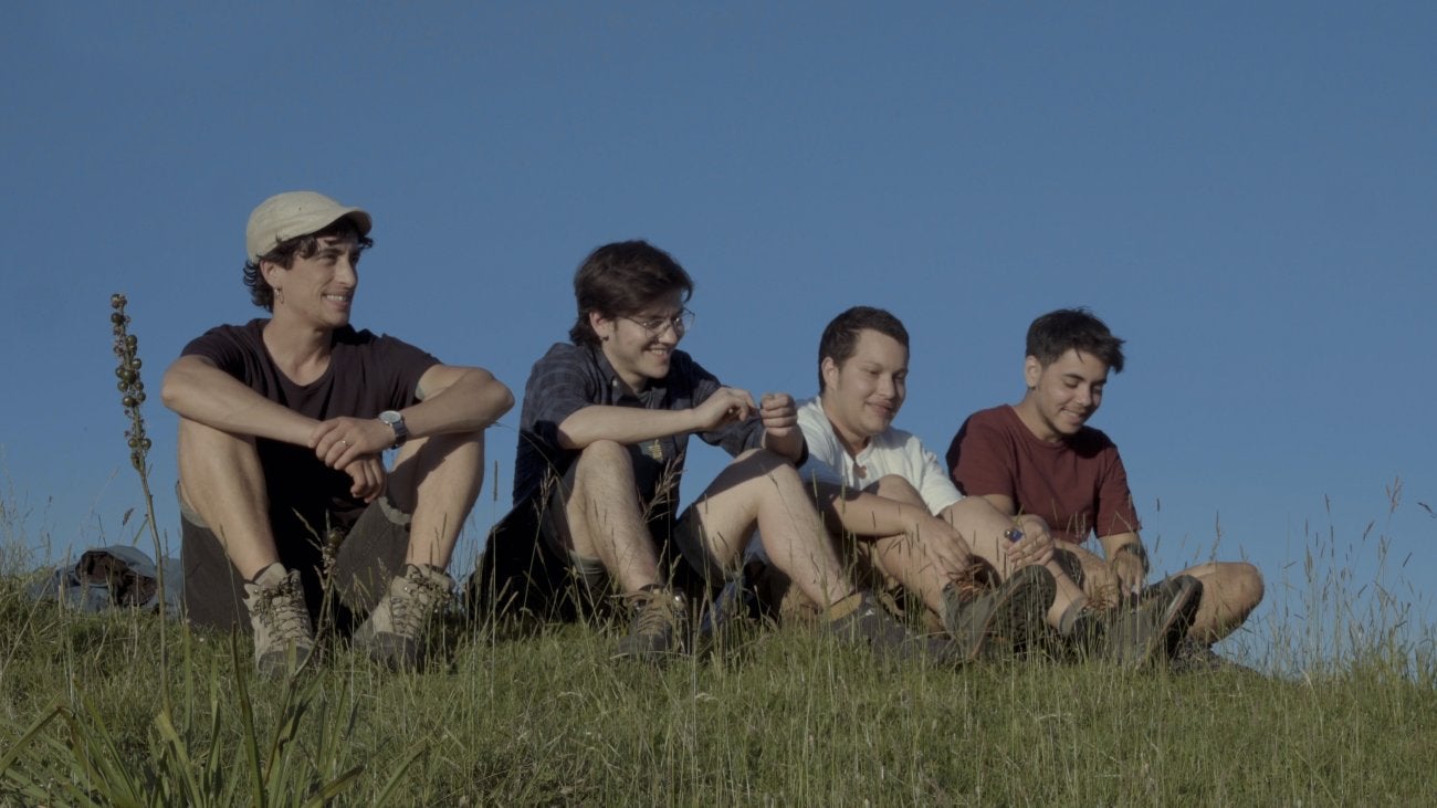 Image of four Trans masc film participants, young Italian men sitting on a hill and smiling after a hike. Blue sky is behind them.