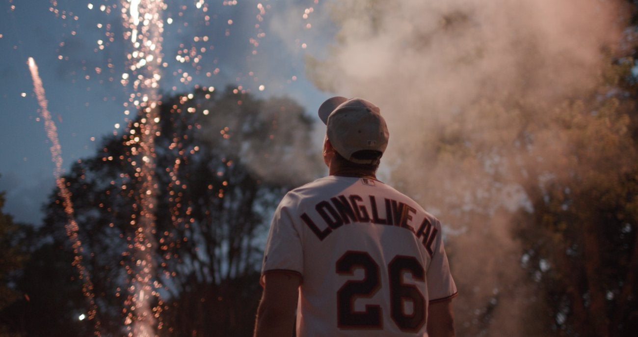Image of a man in a basketball cap wearing a shirt that says along the back "Long Live Al" Looking up at fireworks. 