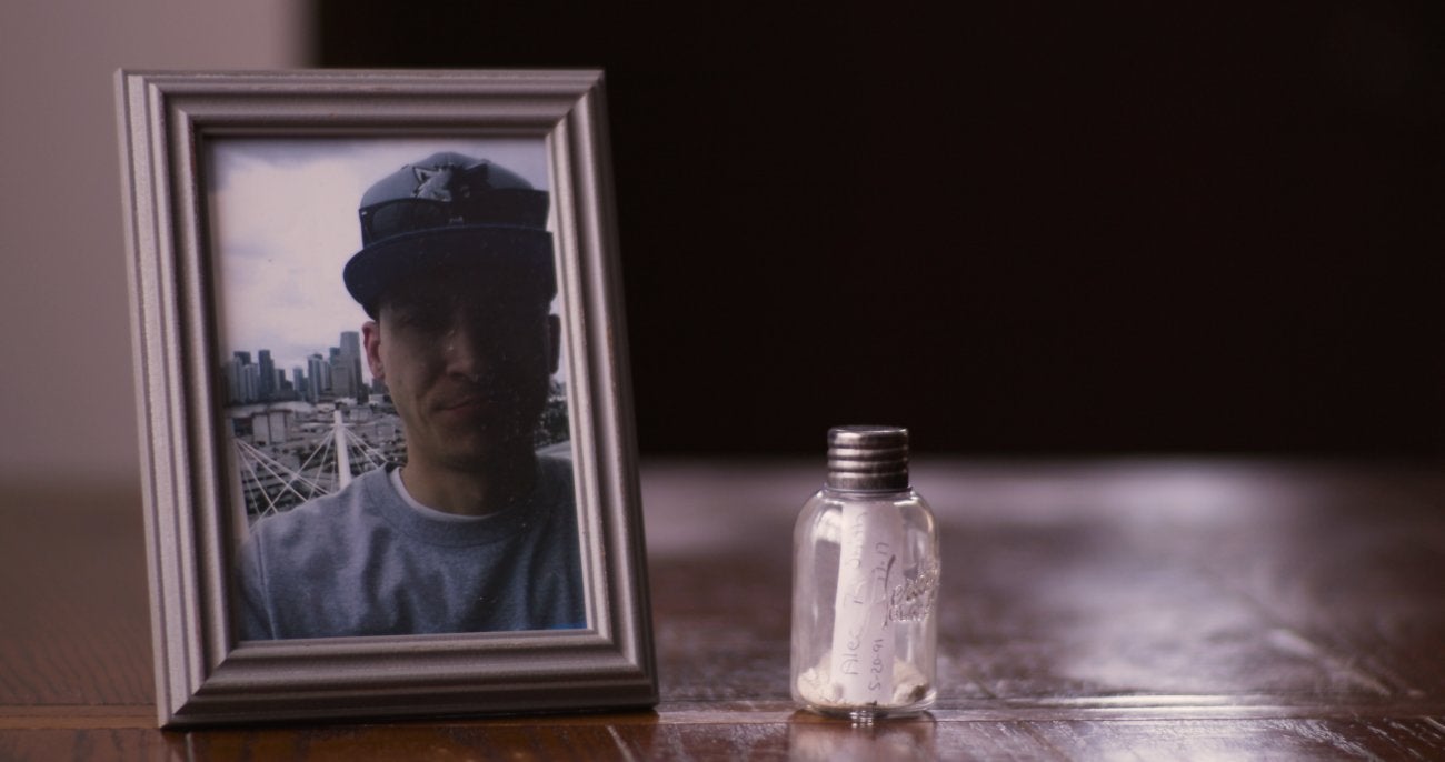 Photo of a young man in a picture frame sitting on a brown table. Next to picture frame sits a vial with a note inside. 