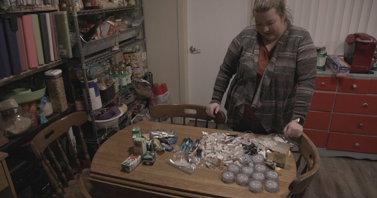 Image of white woman looking down at a table which is covered with medical supplies. She stands to right of a pantry cabinet full of home goods. 