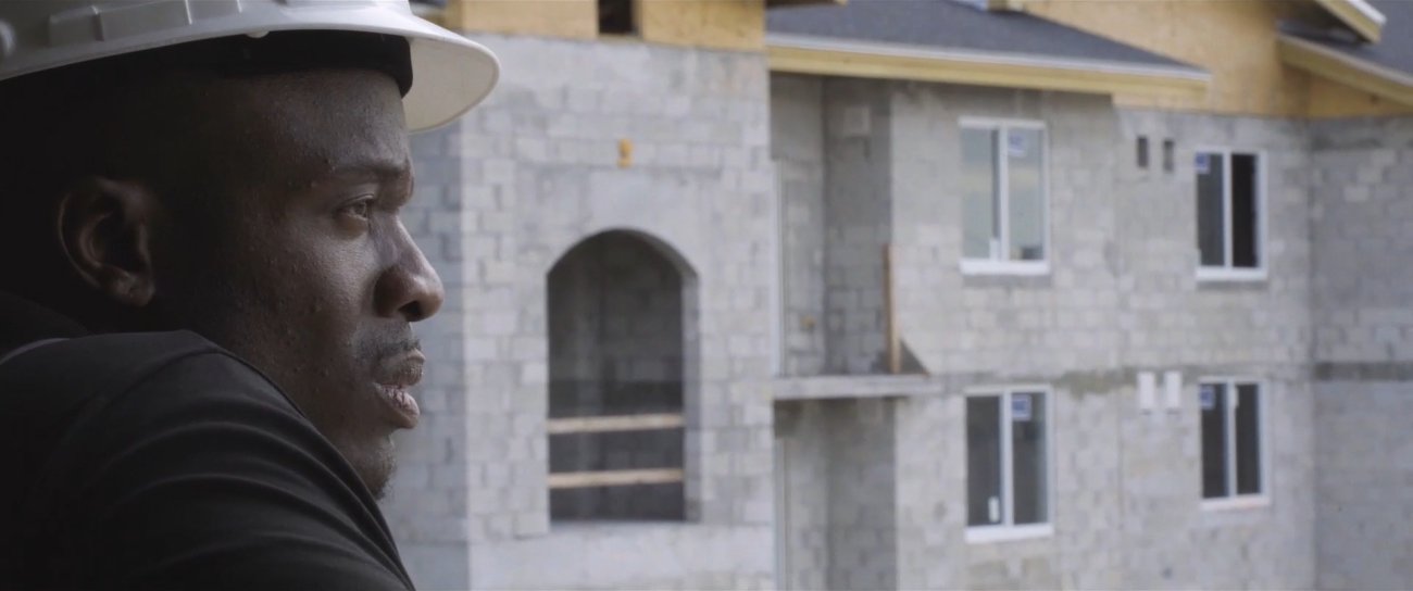 Image of Black man wearing a hard hat looking towards the right. He stands in front of a construction site where a building with detailed arches is being built. 