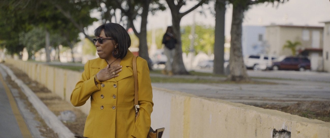 Photo of a Black woman wearing a yellow trench coat. She has a pocketbook on one shoulder and raises her left hand, holding it to her chest. She wears sunglasses and an expression filled with emotion. She is standing in what looks like a park with a sidewalk. 