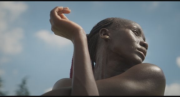 African woman looks to the right, braids of hair close to her head. Her arm stretches out to the left in front of a blue sky.