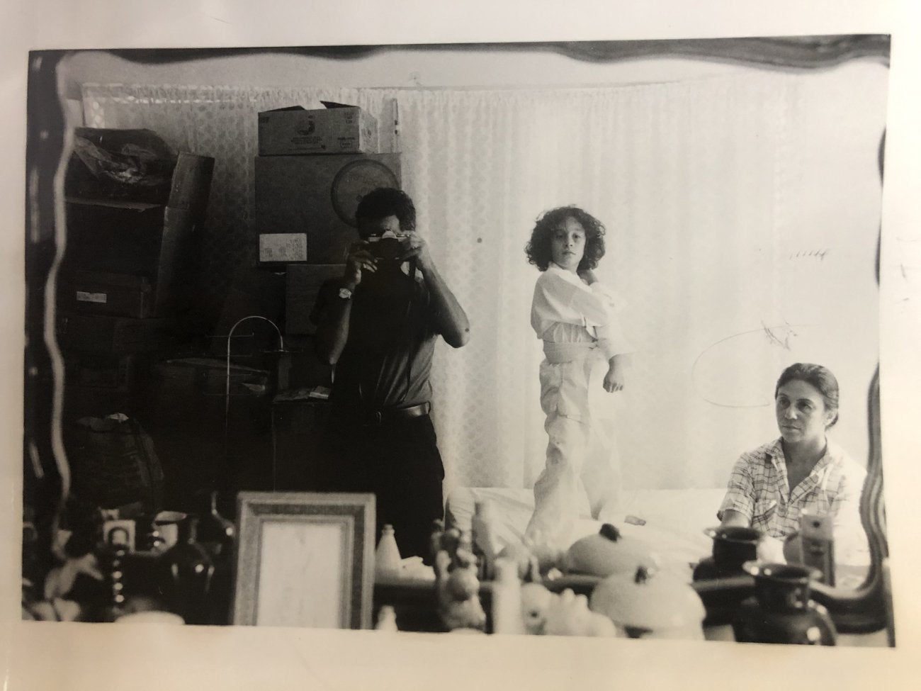 A B&W archival shot of a male figure in a photography studio taking self-portrait in a mirror, with a woman posing in the background.