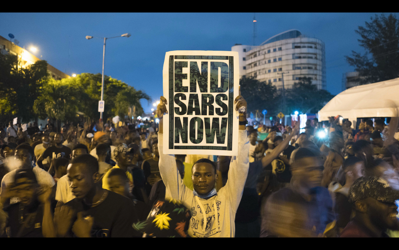 A crowd with central male holding a sign that reads END SARS NOW