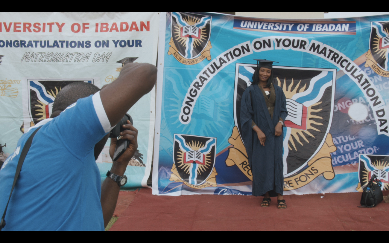 A photographer takes a picture of a student in cap and gown in front of a school banner