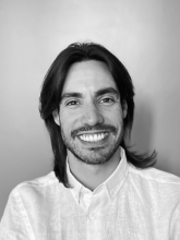 Image of non-binary person smiling with long hair, mustache and white button down shirt.