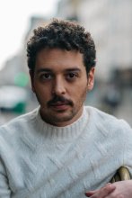 Man with dark curly hair and a moustache looks at the camera