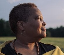 Image of Black woman looking towards the right face towards the sun. She has short brown and gray hair and stands with a field of grass and blue sky behind her.