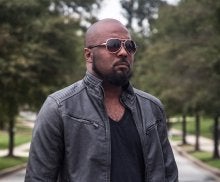 Man with bald head and facial hair wearing leather jacket and sunglasses