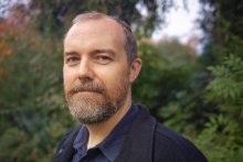 Image of man with brown beard standing in front of greenery